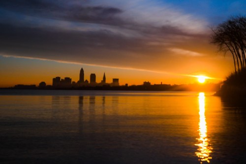 Sunset over a city skyline reflected in water, with silhouettes of buildings and trees against a colorful sky.