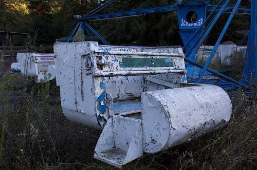 An old, weathered amusement park ride seat sits abandoned among overgrown grass and rusting machinery.