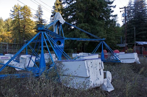 An abandoned Ferris wheel with rusted blue structure and white gondolas, surrounded by overgrown vegetation and trees.
