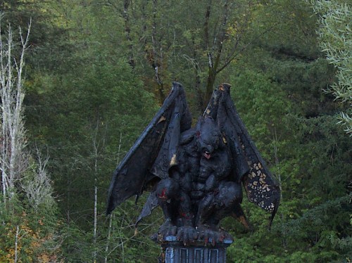 A dark, winged gargoyle statue perched on a pedestal, surrounded by lush green trees.