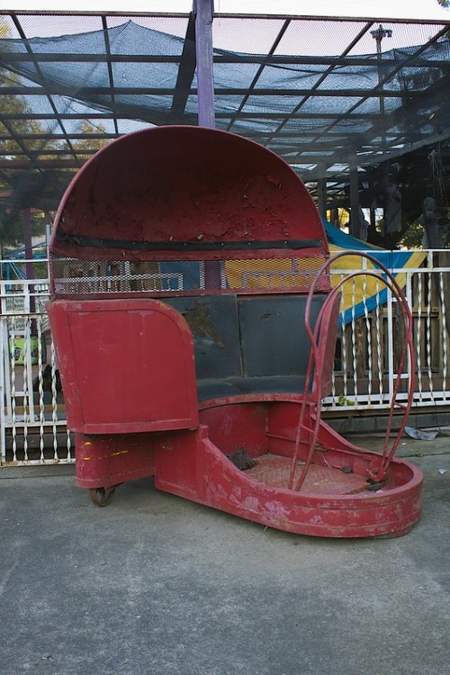 A worn, red amusement park ride seat sits on a concrete surface, surrounded by a fenced area.