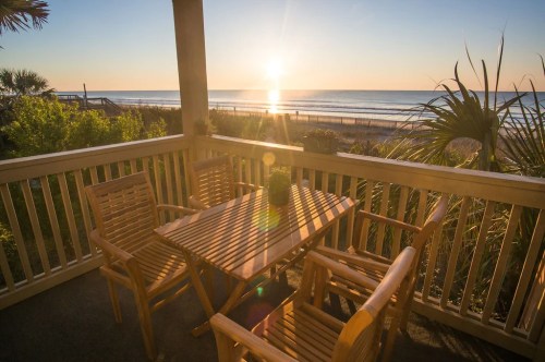 A wooden patio set with four chairs overlooks a beach at sunset, with the sun reflecting on the water.