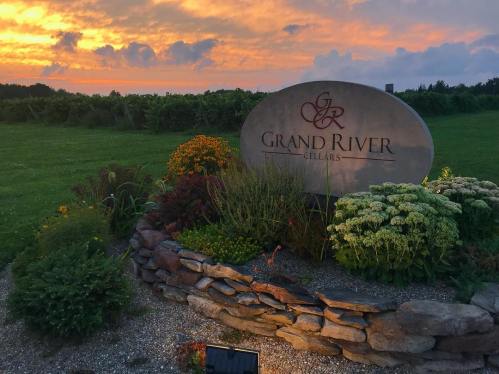 A stone sign for Grand River with colorful flowers and a sunset in the background, set in a green landscape.