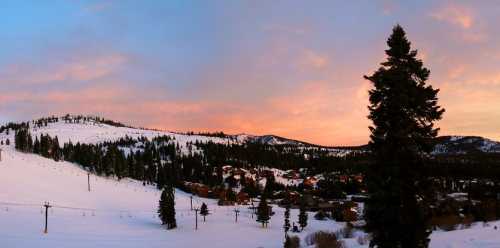 A snowy landscape at sunset, featuring hills, pine trees, and a small town nestled in the valley.