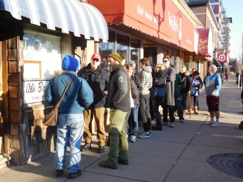 A line of people waiting outside a shop on a sunny day, with storefronts visible in the background.