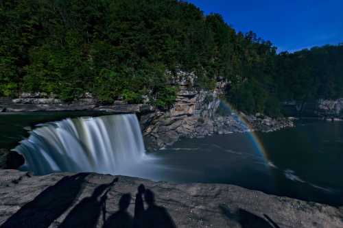 A waterfall cascades into a river, with a rainbow forming in the mist, surrounded by lush green trees and a clear blue sky.