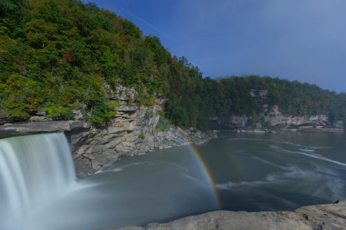 A serene waterfall cascades into a river, with lush green cliffs and a rainbow arching over the scene.