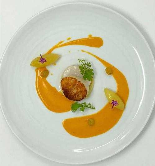 A beautifully plated dish featuring a seared scallop, garnished with sauce, herbs, and decorative elements.