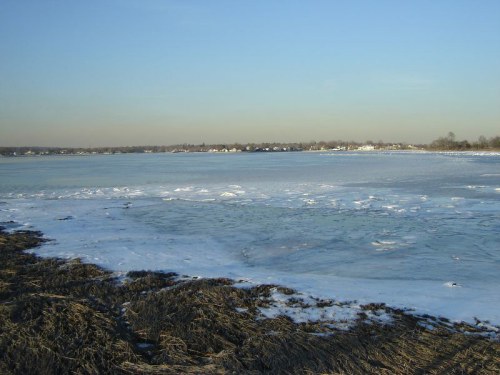 A serene winter landscape featuring a frozen lake, with patches of ice and a clear blue sky in the background.