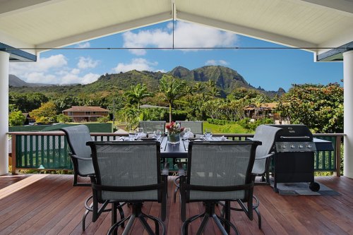 A scenic outdoor dining area with a table, chairs, and a grill, overlooking lush mountains and greenery.
