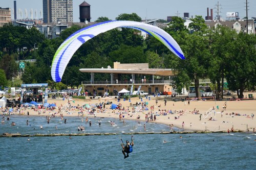 A paraglider soars above a busy beach with people enjoying the sun and water activities below.