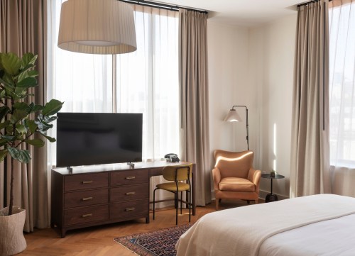A modern hotel room featuring a bed, a TV on a wooden dresser, an armchair, and large windows with sheer curtains.