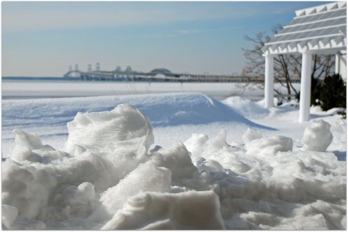 Snow-covered landscape with a distant bridge and a white gazebo, under a clear blue sky.