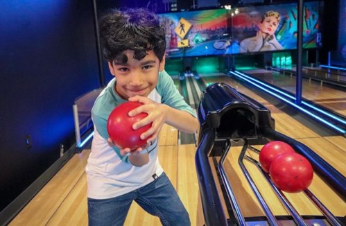 A young boy smiles while holding a red bowling ball, standing near a bowling lane with colorful lights in the background.