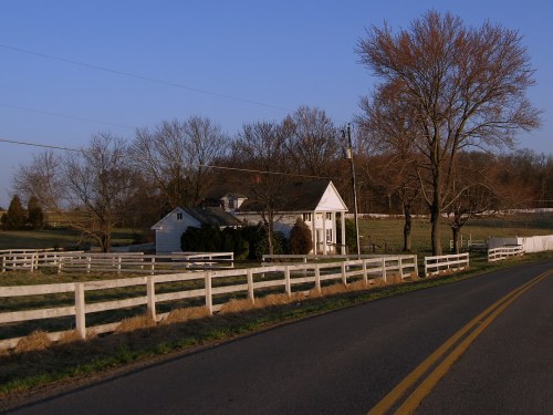 A quaint white house beside a road, surrounded by trees and a white fence under a clear blue sky.