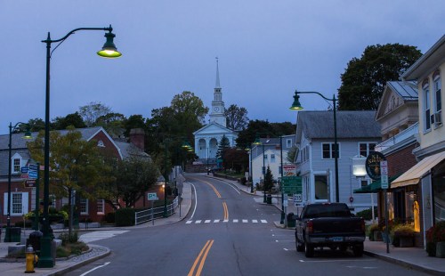 Quiet street scene at dusk, featuring a church steeple in the distance and charming shops lining the road.