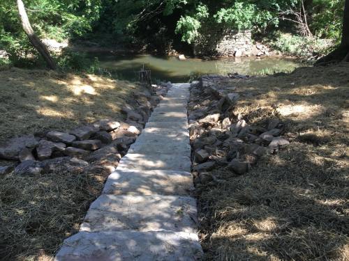 A stone pathway leads down to a calm stream, surrounded by trees and grassy areas.