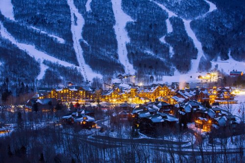 A snowy mountain village illuminated at night, with ski slopes visible in the background.