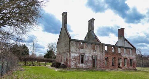 Ruins of a brick building with two tall chimneys, surrounded by grass and trees under a cloudy sky.