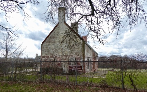 A weathered brick building behind a fence, surrounded by bare trees and cloudy skies. A sign is visible in front.