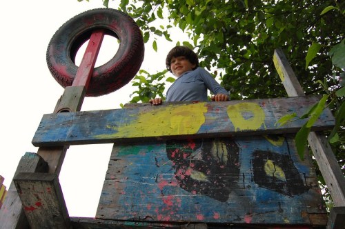 A child stands on a colorful wooden play structure, looking down from a raised platform surrounded by greenery.