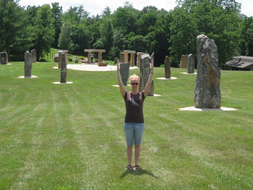 A person stands with arms raised in a grassy area surrounded by large stone structures and trees in the background.