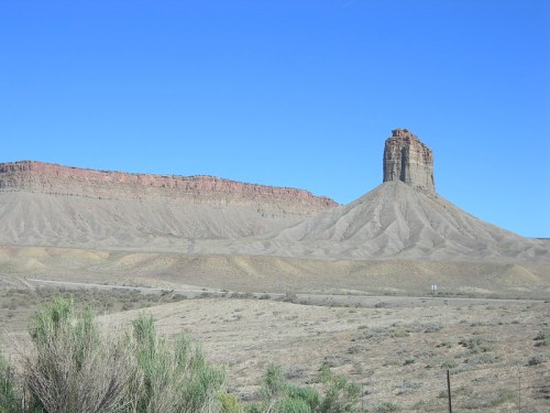 A tall rock formation rises from a dry, barren landscape under a clear blue sky. Sparse vegetation is visible in the foreground.