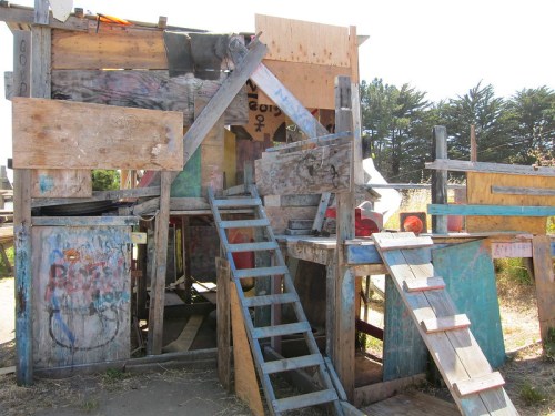 A colorful wooden play structure made of various materials, featuring stairs and multiple levels in an outdoor setting.