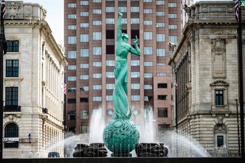 A green statue of a woman rising from a globe, surrounded by a fountain, with buildings in the background.