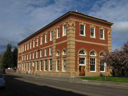 Historic brick building with large windows, decorative arches, and flags, set against a cloudy sky.