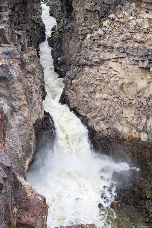 A rushing river flows through a narrow canyon, surrounded by steep, rocky cliffs. Mist rises from the water.
