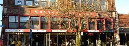 A brick building with large windows and a tree in front, featuring shops and signs for various businesses.