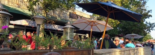 A bustling outdoor café scene with people seated under umbrellas, surrounded by greenery and warm sunlight.