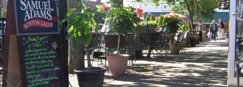Outdoor dining area with tables, greenery, and a sign for Samuel Adams Boston Lager. People are walking in the background.