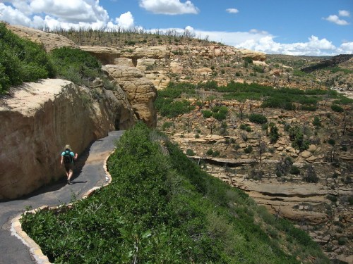 A hiker walks along a winding path through a rocky landscape with green vegetation and blue skies above.