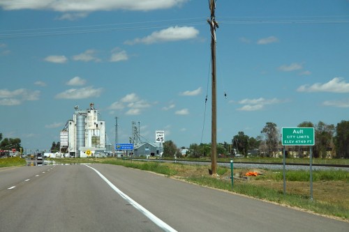 A highway scene showing a sign for Ault city limits, with silos and a clear blue sky in the background.