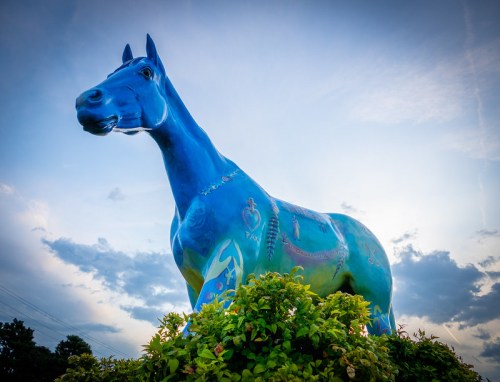 A large blue horse statue adorned with colorful designs, standing amidst green foliage under a cloudy sky.