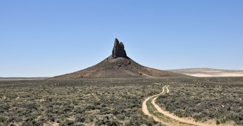 A tall, rocky formation rises from a flat, arid landscape, with a dirt path winding through the surrounding vegetation.