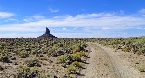 A dirt road leads through a desert landscape to a prominent rock formation under a blue sky with scattered clouds.