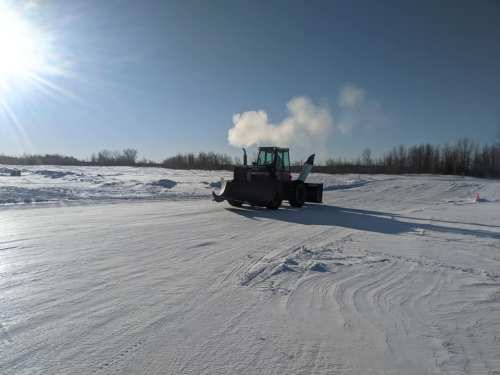 A snowplow clears a snowy landscape under a clear blue sky, with sunlight shining in the background.