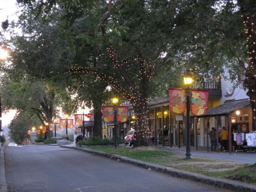 A charming street lined with trees and colorful banners, illuminated by street lamps during twilight.