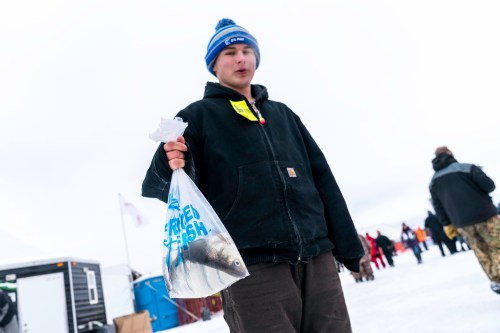 A person in a winter coat holds a bag of fish, standing on snow with people in the background.