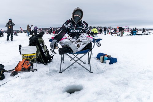 A person sitting on a chair on ice, fishing through a hole, surrounded by others in a snowy landscape.
