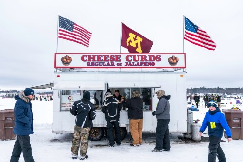 A food truck selling cheese curds, with people in winter clothing and flags overhead, set in a snowy landscape.