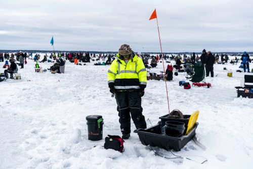 A person in a bright jacket stands on a snowy ice fishing site, surrounded by other anglers and fishing gear.
