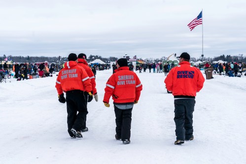 Three members of a sheriff dive team walk on snow, with an American flag and a crowd in the background.