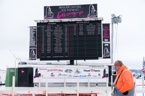 A scoreboard displaying results for an ice fishing event, with a person in the foreground.