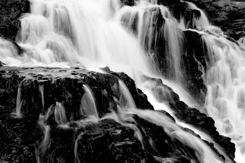 A black and white image of cascading water flowing over dark, rocky surfaces.