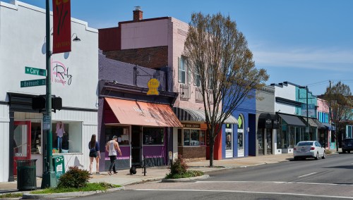 A vibrant street scene with colorful storefronts, trees, and pedestrians on a sunny day.