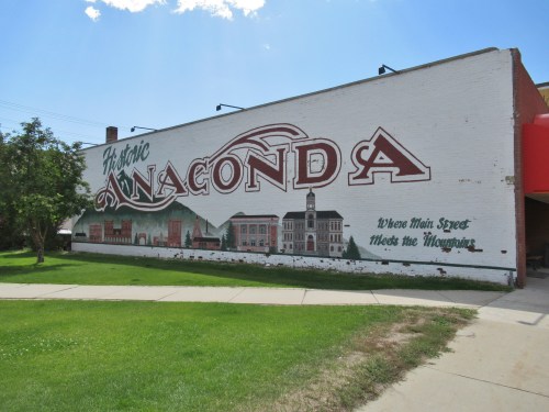 Mural on a brick wall reading "Historic Anaconda" with buildings and mountains depicted, surrounded by green grass.